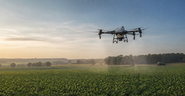 DJI Agras T50 agricultural spraying drone above a crop field with a ground boom sprayer in the distance